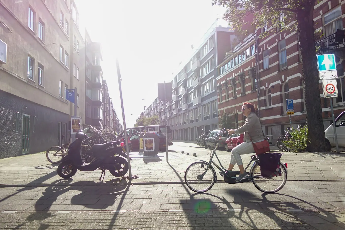 A woman with pannier bags cycling down a sunny street lined with brick buildings