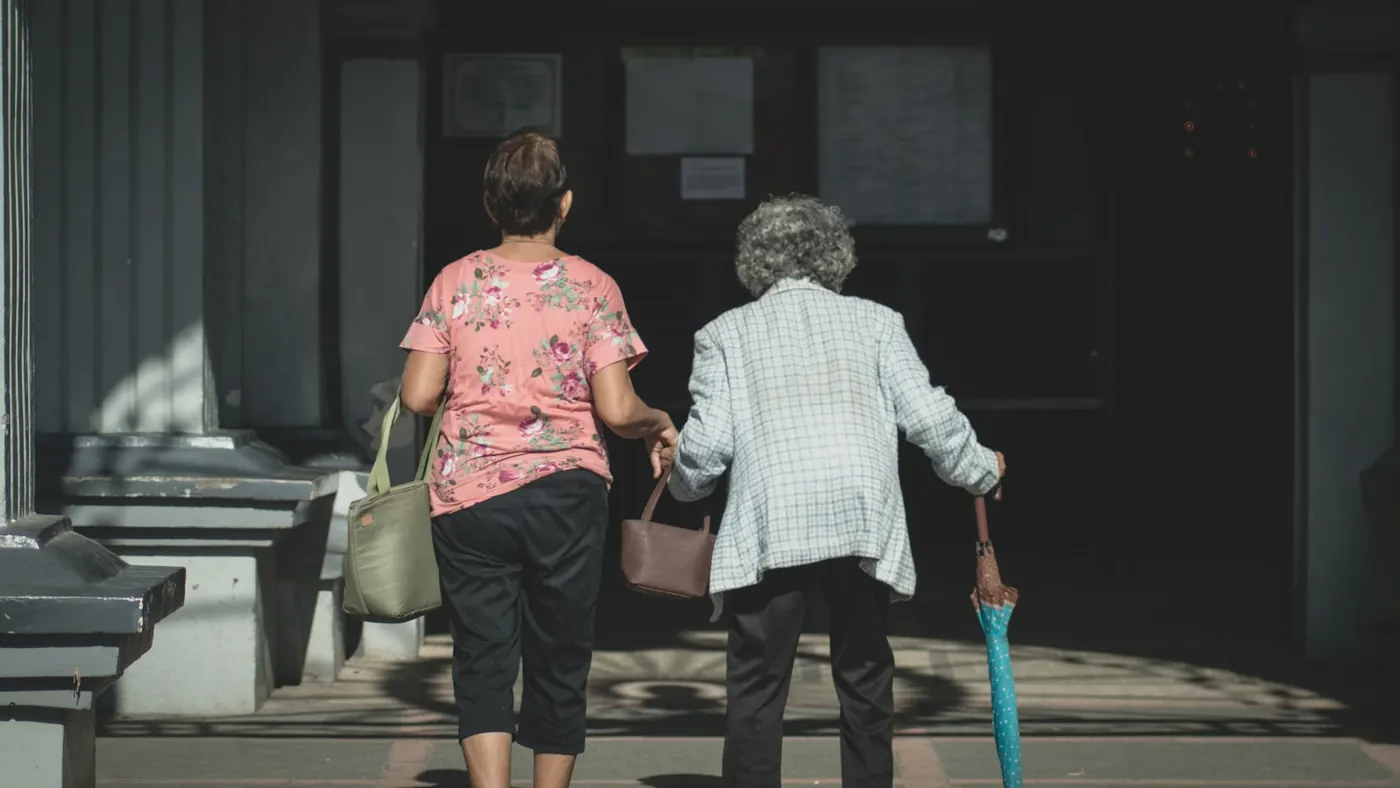 A woman walking arm in arm with an older woman holding an umbrella, in the sunshine